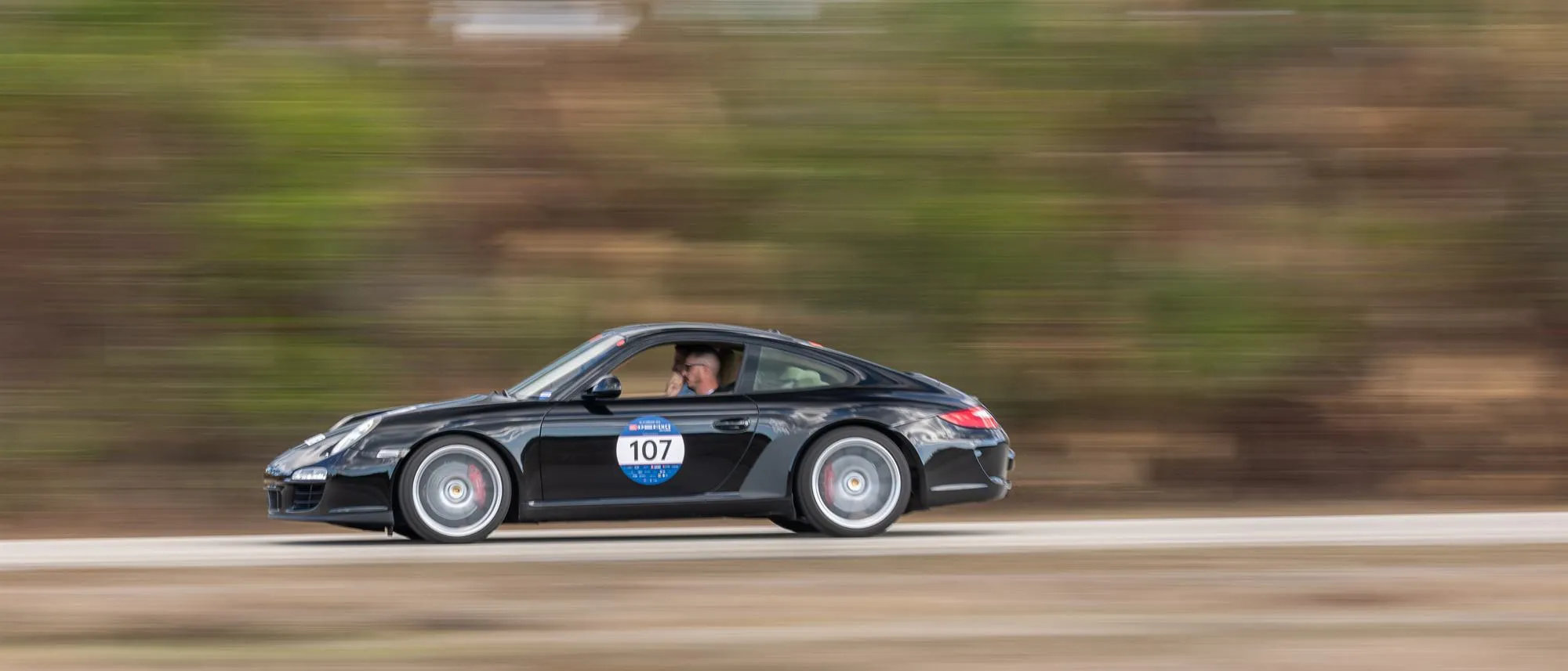 Car 107, a black 2010 Porsche 911 Carrera S driven by Erick Mahle, at speed on a Florida stage of the 1000 Miglia Experience USA, February 2026